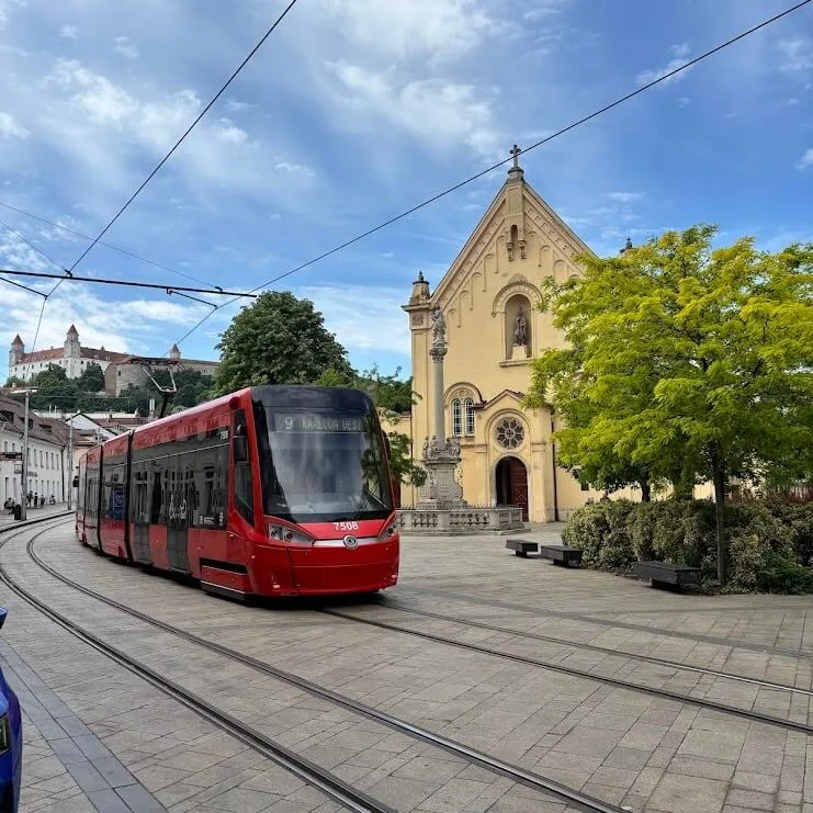Eine rote Straßenbahn fährt an einer gelben Barockkirche in Bratislava vorbei, mit der Burg am Horizont im Hintergrund.