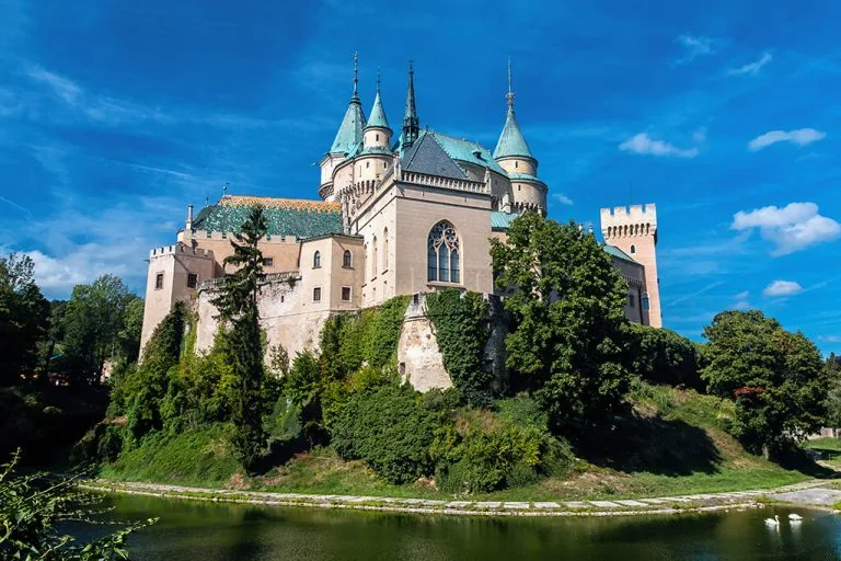 Das Schloss Bojnice in der Slowakei, dessen Türme über einem bewaldeten Hügel und dem Wassergraben aufragen.
