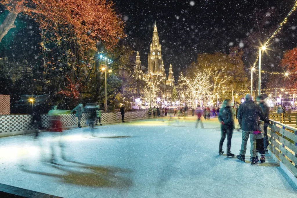 Schlittschuhläufer auf einer beleuchteten Freiluft-Eisbahn im Schneefall, dahinter das angestrahlte Wiener Rathaus.