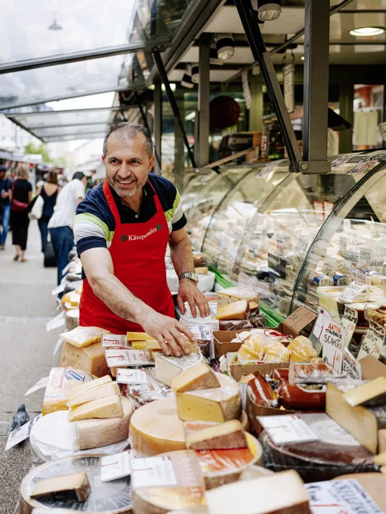 A smiling cheesemonger at a stall in the Naschmarkt in Vienna, behind a counter of whole and cut cheeses.
