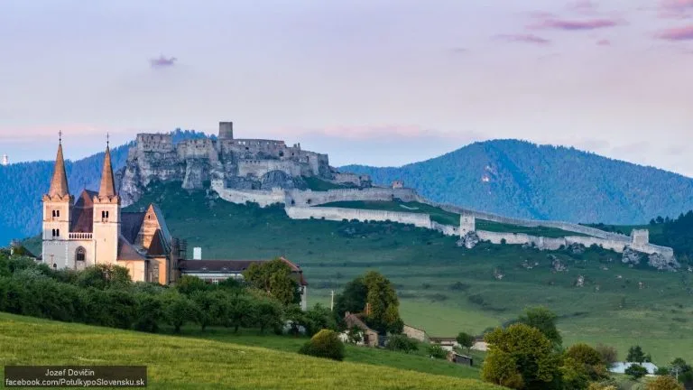 Spiš Castle at dusk, seen across the fields from Spišská Kapitula, with the twin towers of St Martin's Cathedral in the foreground.
