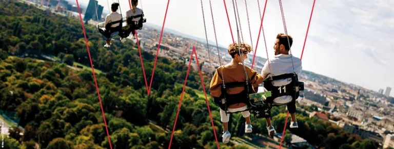 Riders on a high chain-swing ride at the Prater, swinging out above the trees with the rooftops of Vienna below.