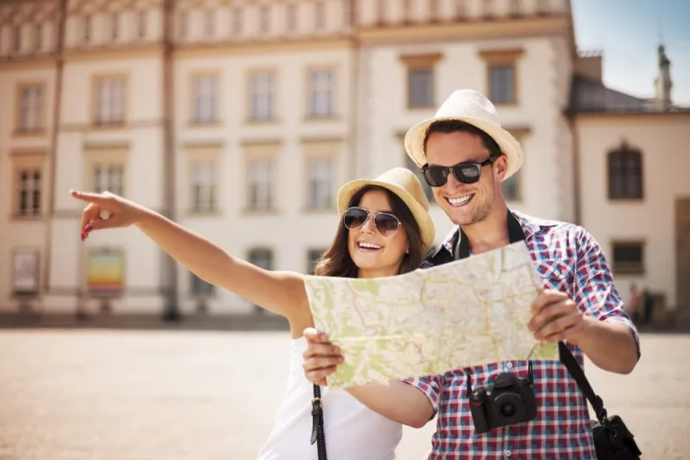 Two travellers in sun hats with a folded map, one of them pointing, in front of a historic town building.