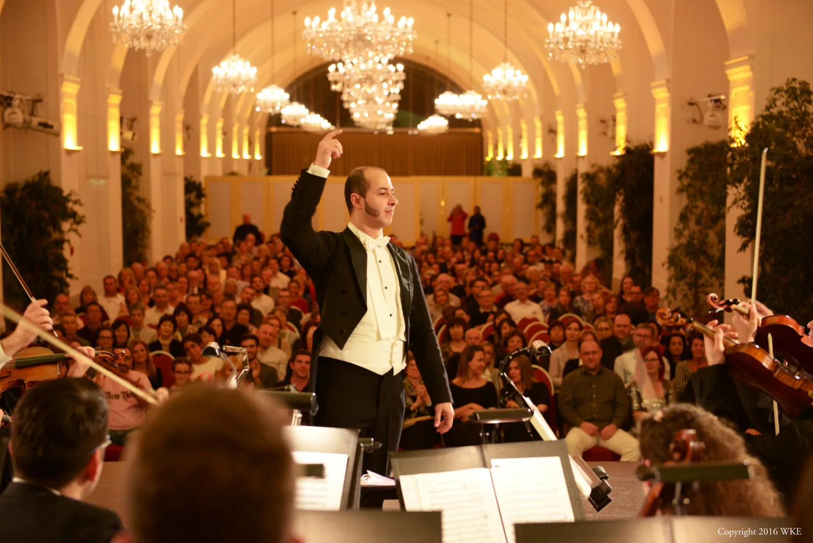Ein Dirigent und ein Orchester bei einem Konzert vor vollem Publikum in einem Saal mit Kronleuchtern.