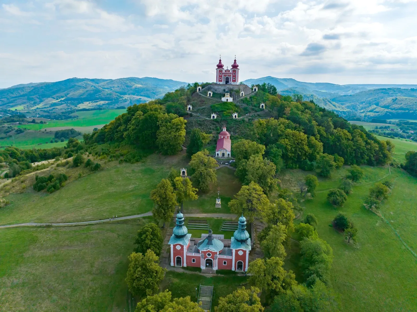 The baroque calvary at Banská Štiavnica from the air, its white chapels climbing the wooded hill to the twin-towered church at the summit.