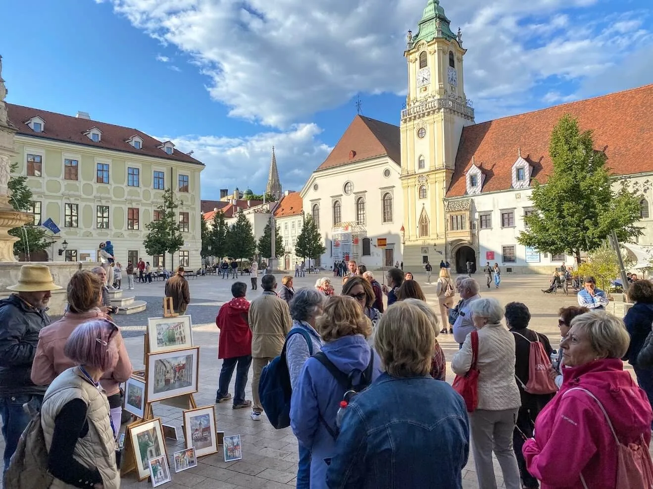 A tour group on Bratislava's Main Square, with watercolour paintings for sale on easels and the tower of the Old Town Hall behind.