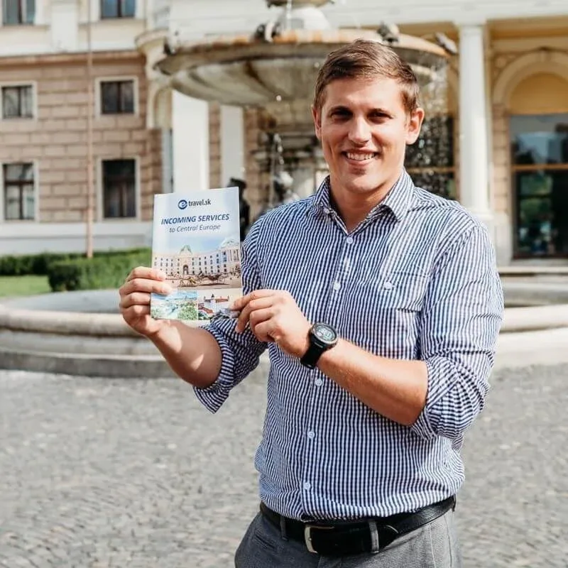Andrej Stanik, VIP guide at E-TRAVEL.SK, holding the company’s Incoming Services to Central Europe brochure beside a fountain.