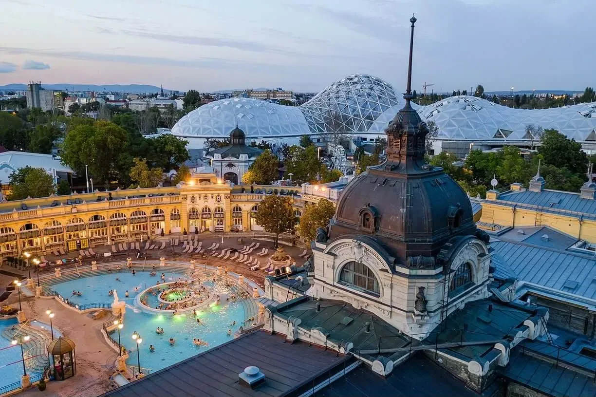 The outdoor thermal pools of the Széchenyi Bath in Budapest at dusk, ringed by the yellow neo-baroque arcades of the bath complex.