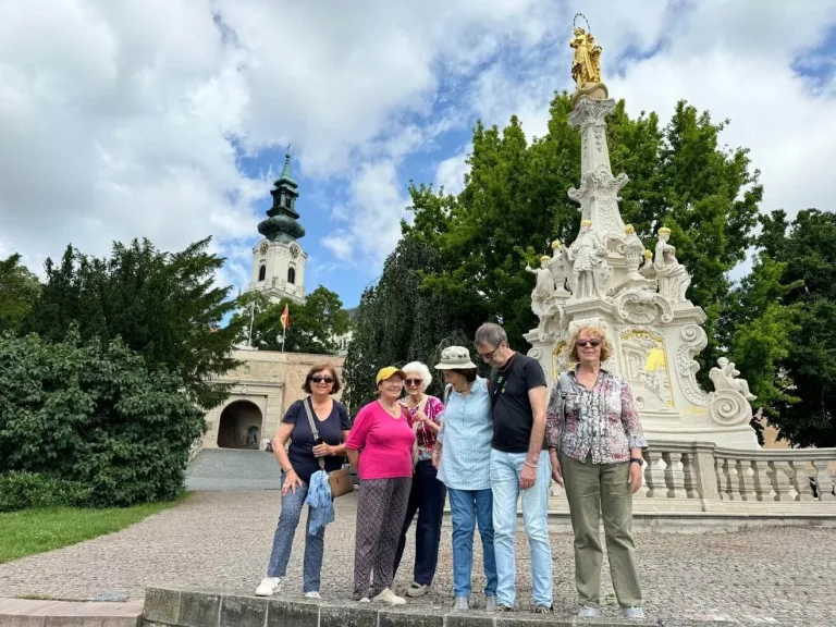 A small group of travellers posing beside the baroque plague column below Nitra Castle in Slovakia, with the castle gateway and the cathedral tower behind them.