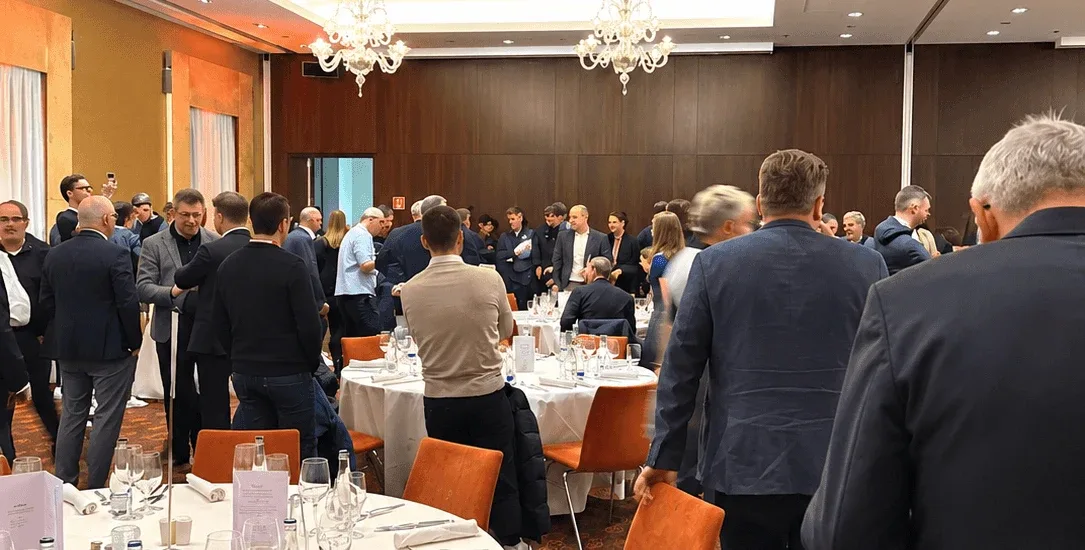 Guests mingling between the dinner tables at a corporate gala dinner in a chandelier-lit hotel ballroom.