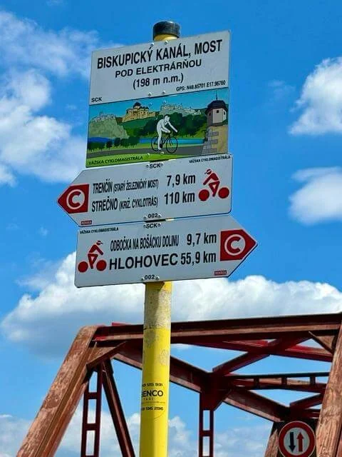 A Váh cycle route signpost at the Biskupický canal bridge, pointing to Trenčín 7.9 km and Hlohovec 55.9 km, with the red steel bridge behind.