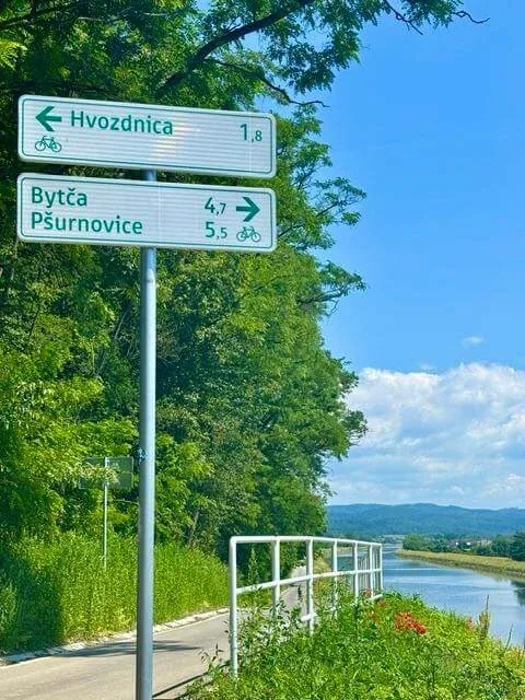 A cycle route signpost to Hvozdnica, Bytča and Pšurnovice beside the Váh canal, with wooded hills behind.