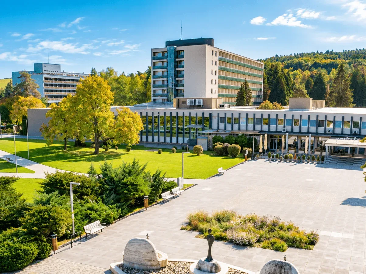 The spa grounds at Dudince in autumn sunshine, with the spa hotels behind the lawns and trees and the paved promenade in front.