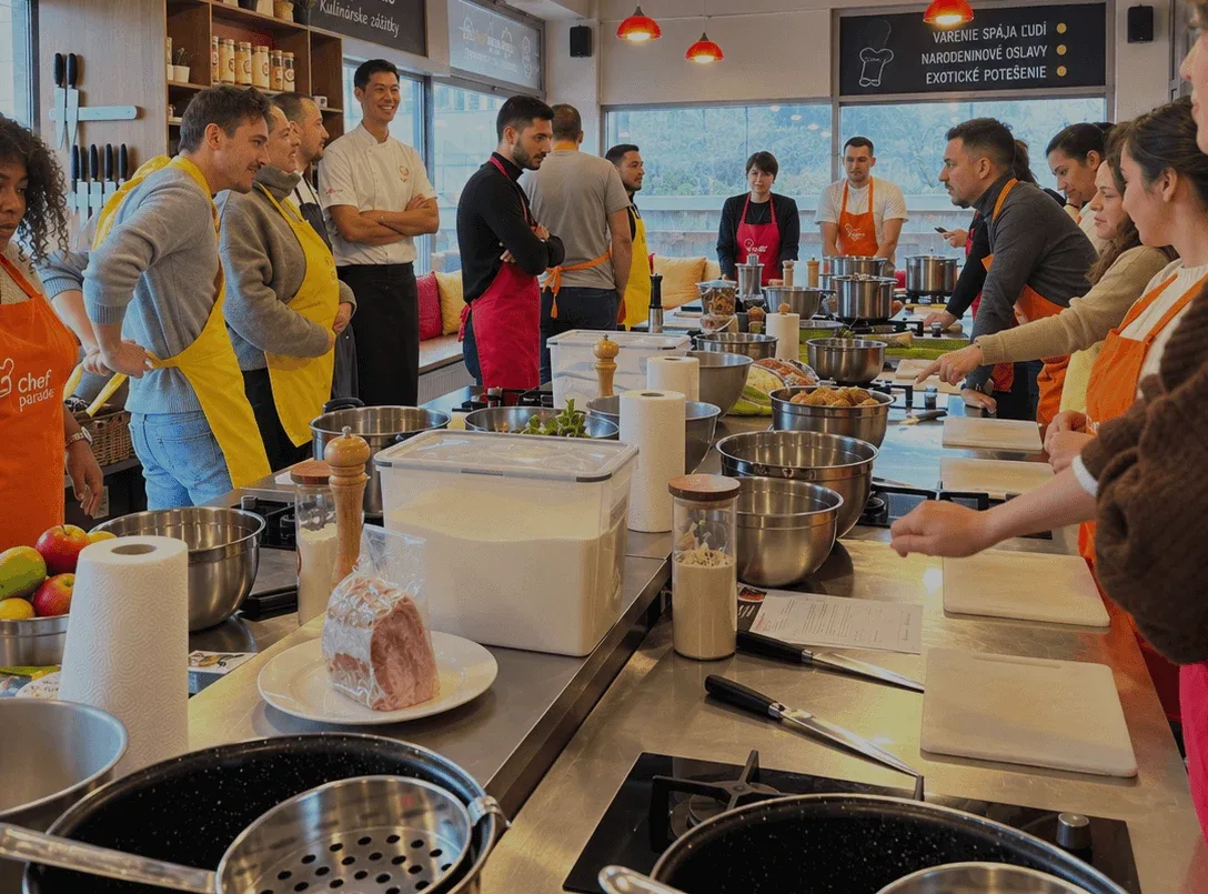 A group cooking class at the Chefparade cooking school, with participants in aprons gathered around the kitchen counters and a chef leading the session.