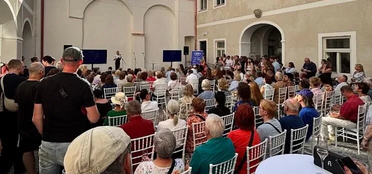 An audience seated in a historic courtyard for an open-air talk, with a speaker on stage between two screens.
