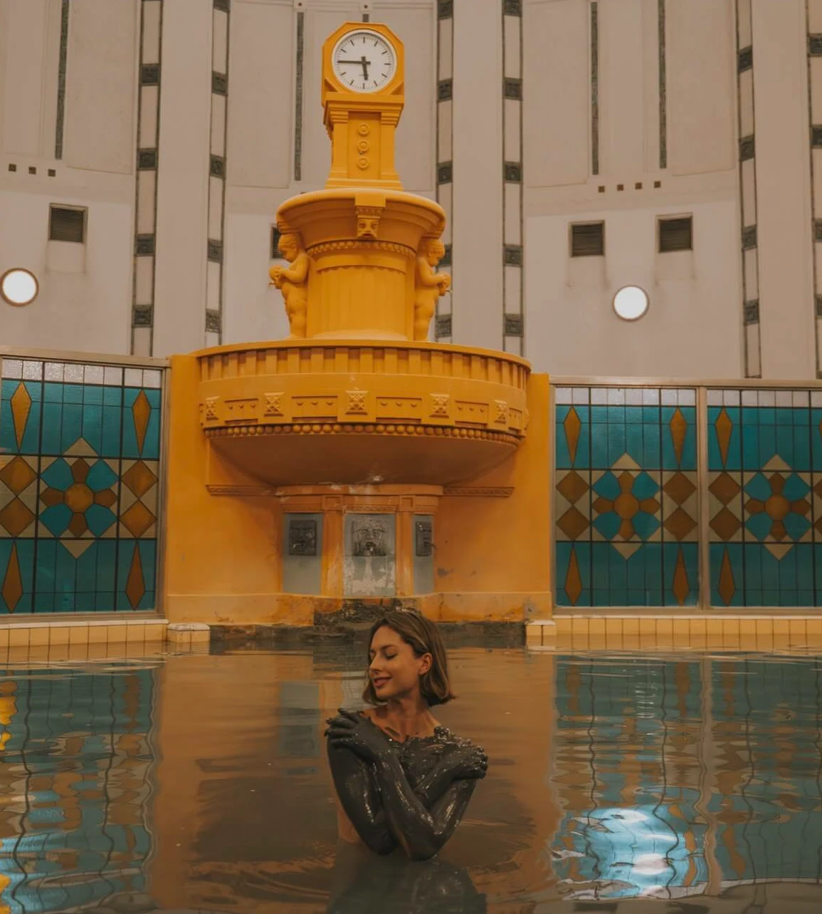 A guest in the thermal mud pool of a historic spa hall in Piešťany, under a domed ceiling with a yellow clock fountain.