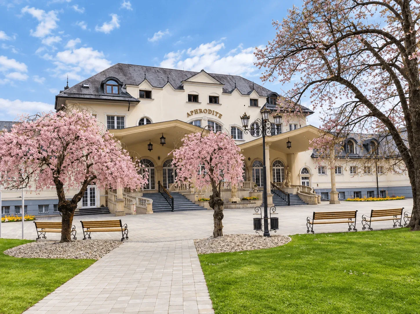 The Aphrodite spa building in Rajecké Teplice, with pink cherry trees in blossom and benches in the spa park in front.