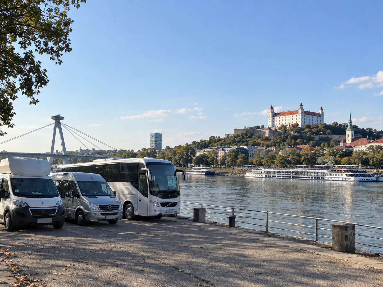 A coach, a minibus and a van parked on the Danube embankment in Bratislava, with river cruise ships moored below Bratislava Castle and Most SNP (the UFO Bridge) in the distance.