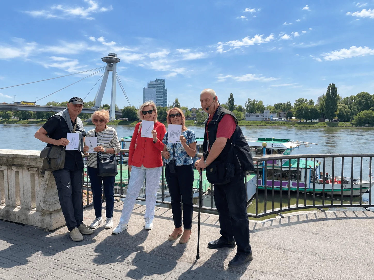 A local guide with a small group on the Danube embankment in Bratislava, with Most SNP (the UFO Bridge) and a moored river boat behind them.
