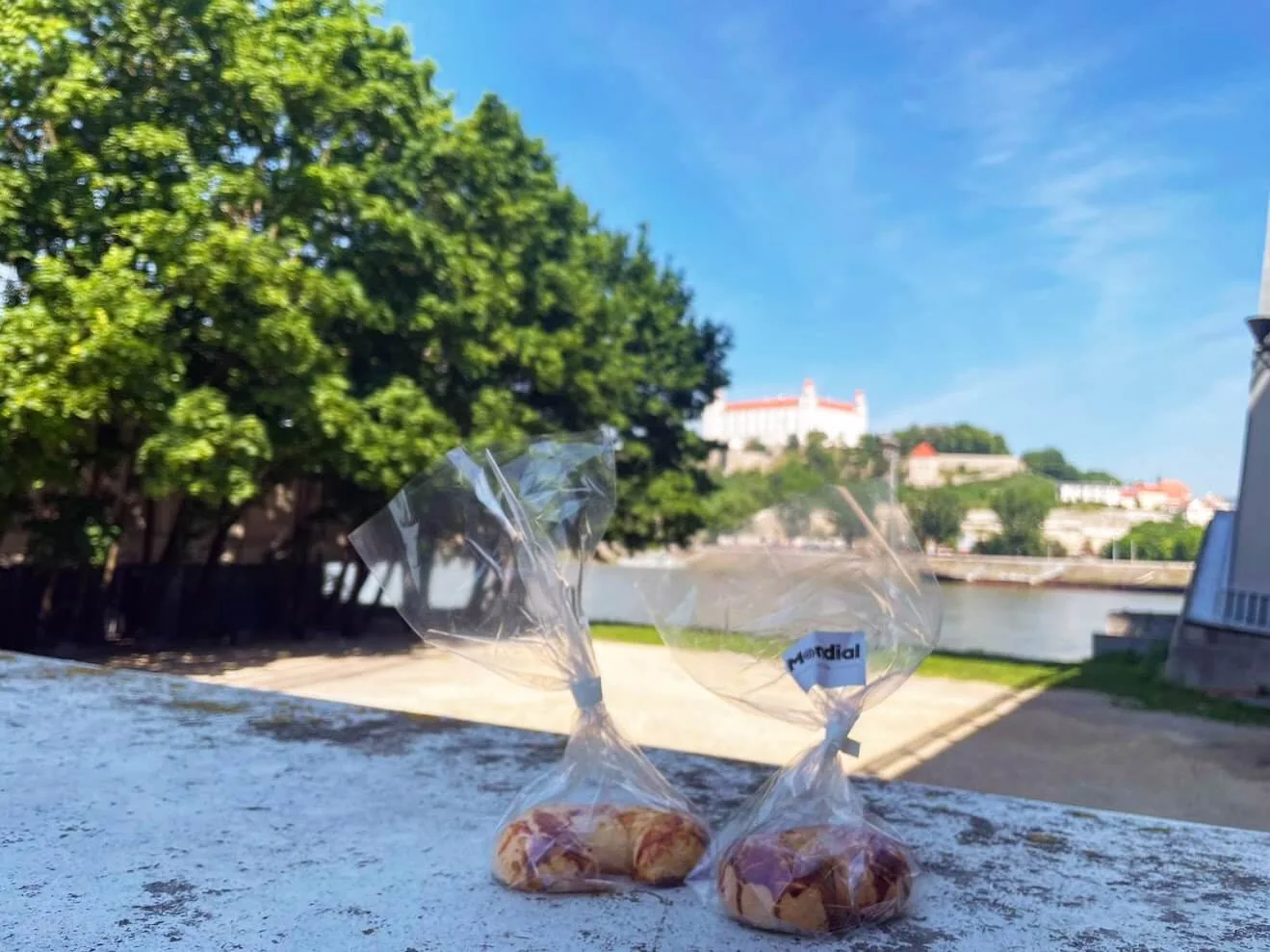 Two wrapped pastries on a wall by the Danube, with Bratislava Castle on its hill across the river.