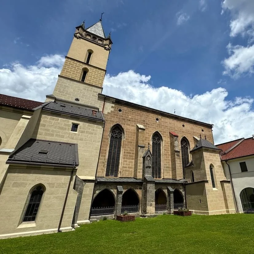The stone Gothic Basilica of St Benedict at Hronský Beňadik abbey, its square bell tower above the nave and a cloister arcade along the lawn.