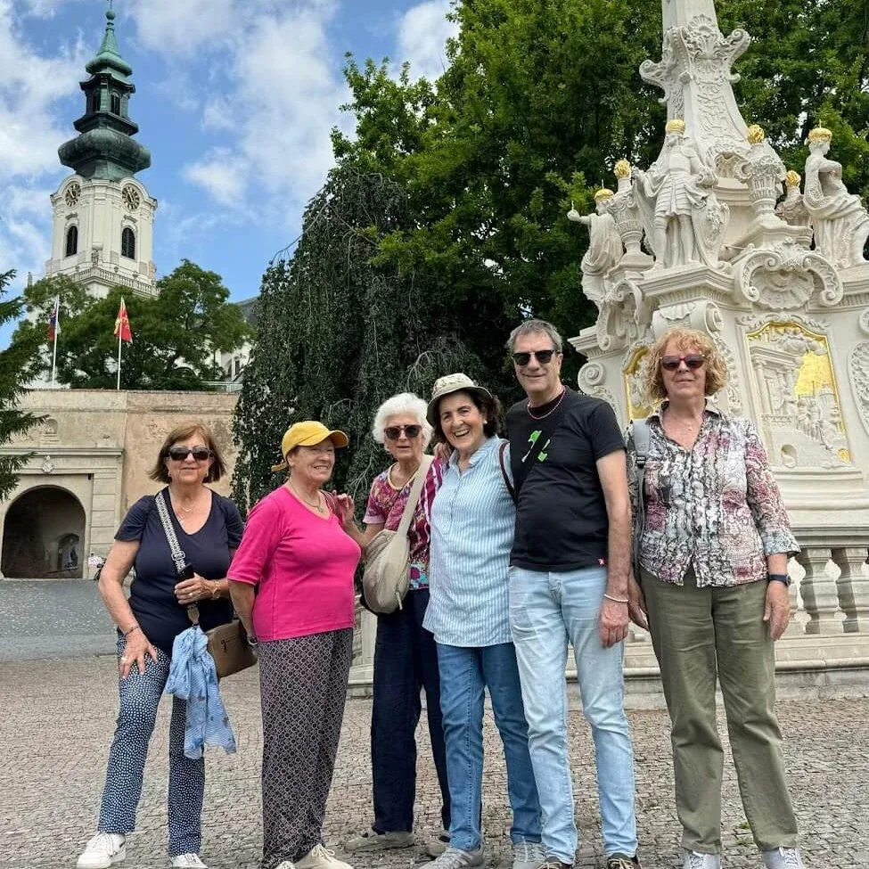 A group of travellers posing beside the baroque plague column on the approach to Nitra Castle, the castle's white clock tower rising behind the trees.