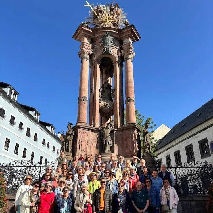 A tour group gathered around a baroque column in a historic town square on a sunny day.