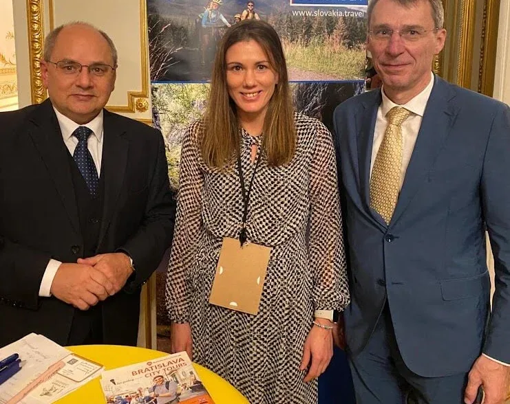 Three people at a Slovakia tourism presentation in a gilded hall, beside a Travel to Slovakia banner and a table of brochures.