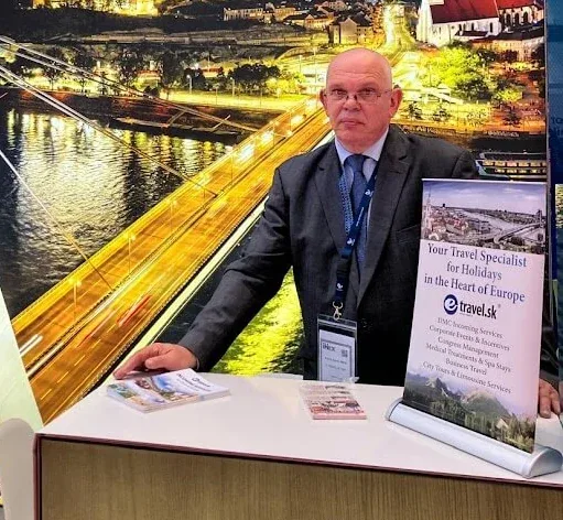 A man at the e-travel.sk stand at a travel trade fair, in front of a large night view of Bratislava and the Danube.