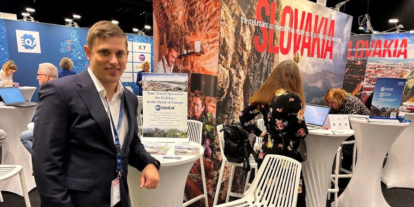 An e-travel.sk representative at the company's stand at a travel trade fair, beside a display board listing its DMC services and a large Slovakia tourism backdrop.