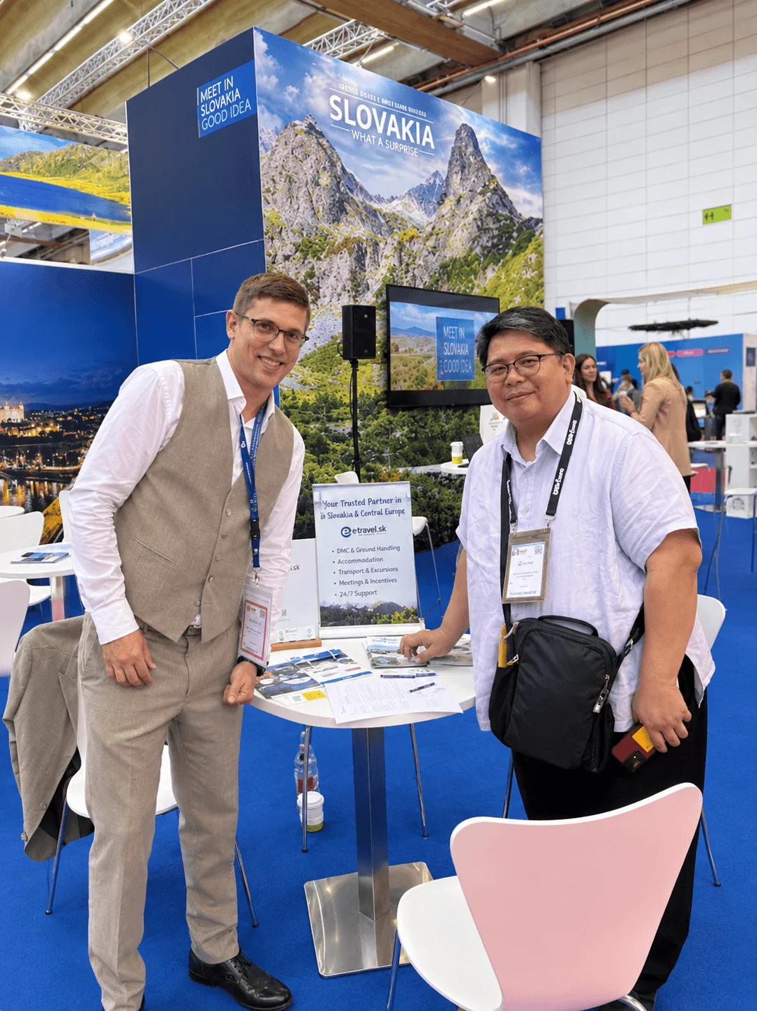 A meeting with a trade partner at the e-travel.sk table on the Slovakia stand at a travel trade fair, in front of the "Slovakia – What a surprise" mountain display.