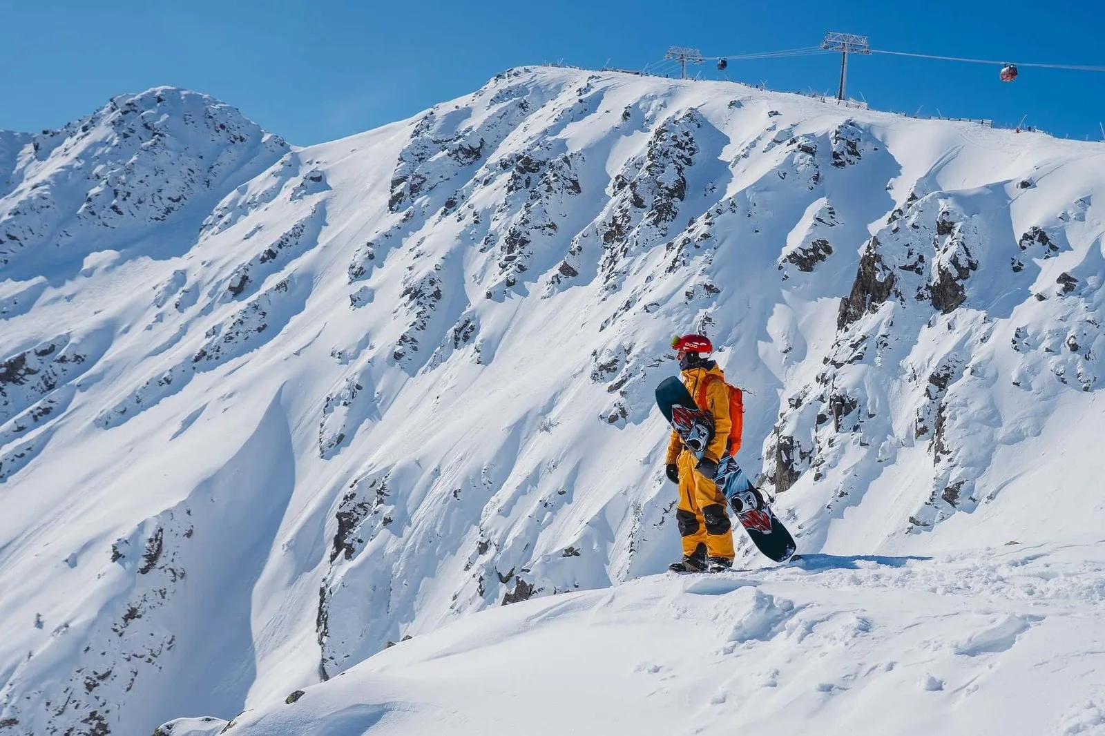A snowboarder standing on a snow-covered ridge at Jasna in Slovakia's Low Tatras, with a gondola crossing the ridge above and snowy peaks beyond.