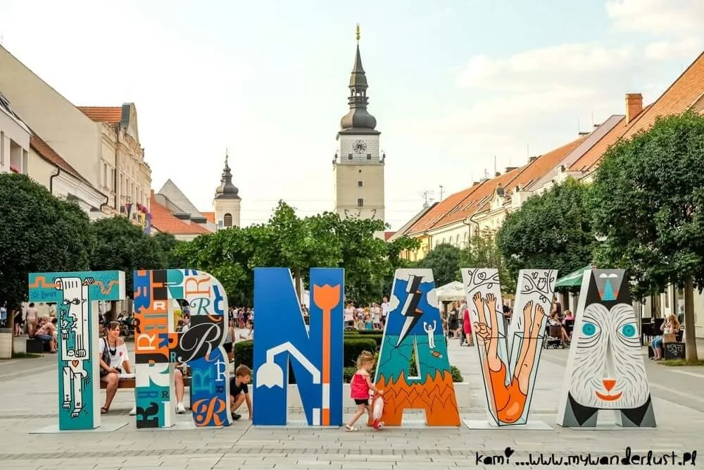 Children playing among the colourful painted TRNAVA letters on the main square, with the town tower and church spires behind.