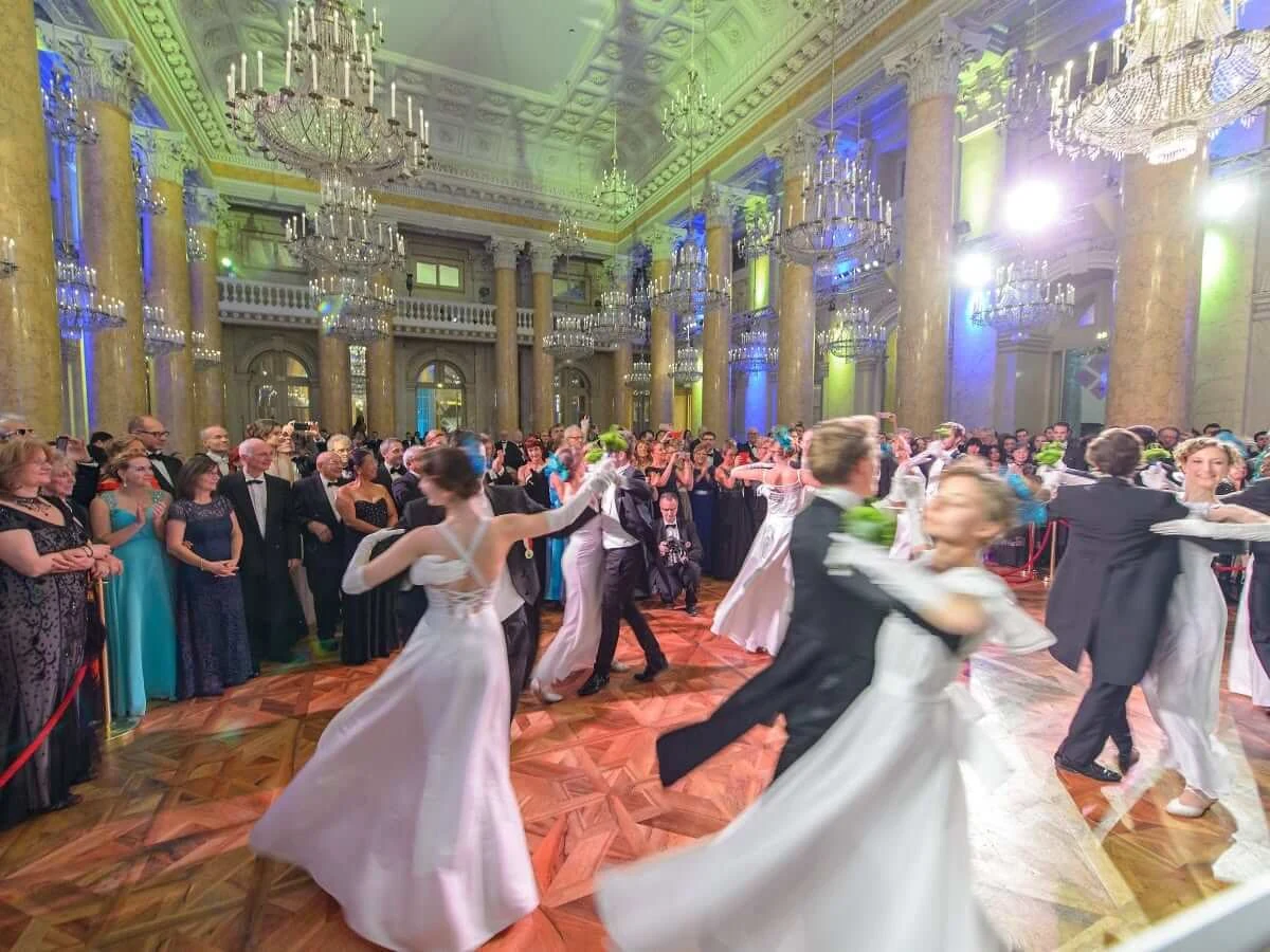 Debutantes in white gowns waltzing with their partners at a Viennese ball, beneath crystal chandeliers in a columned ballroom.