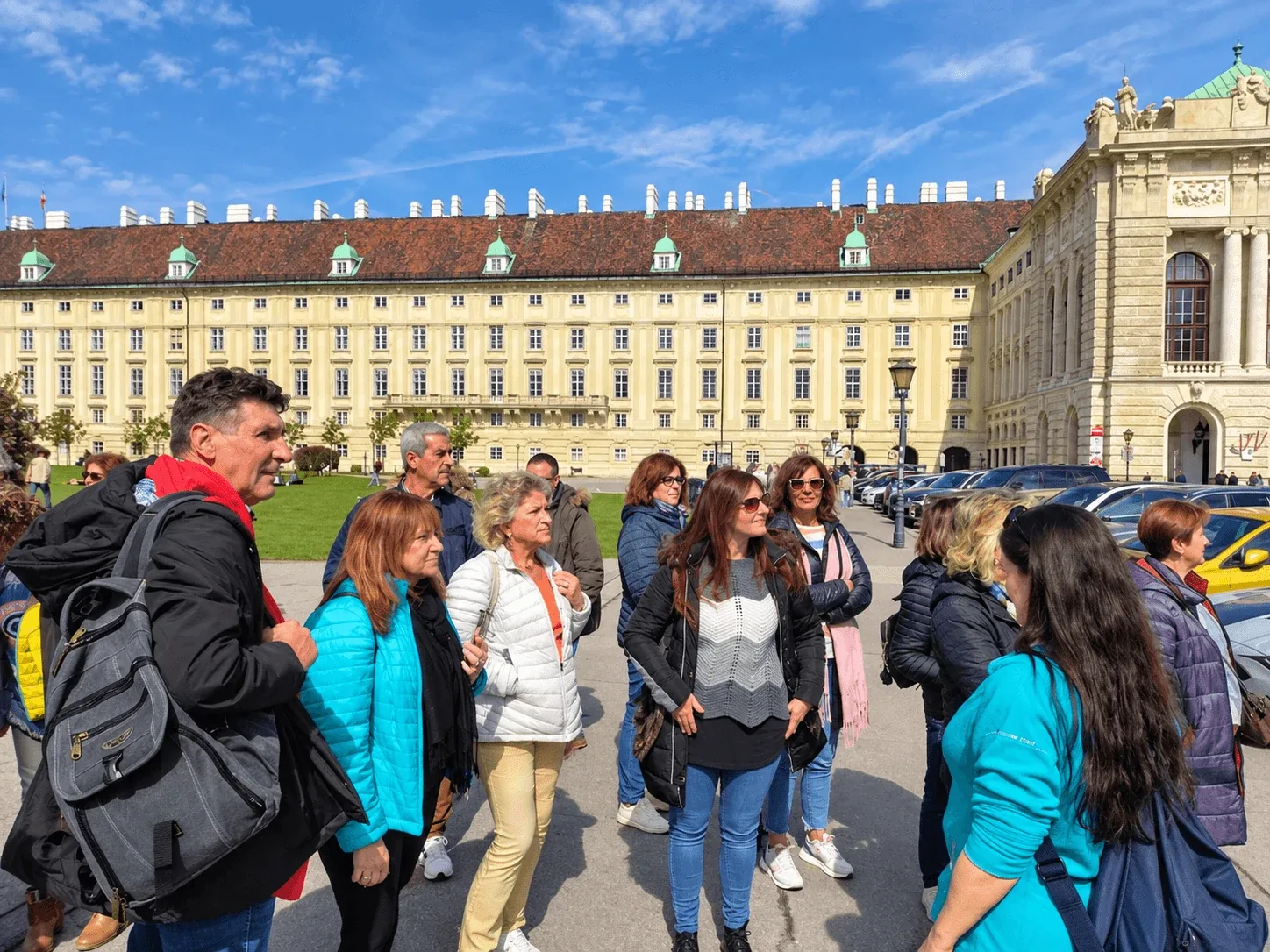 A tour group listening to their guide on Heldenplatz in Vienna, with the long yellow wing of the Hofburg palace behind them.
