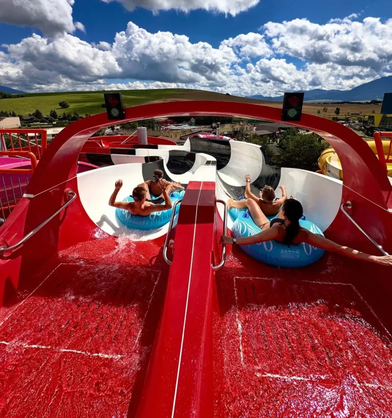 Two pairs of visitors setting off down a red water slide on rubber rings, meadows and mountains beyond the park.
