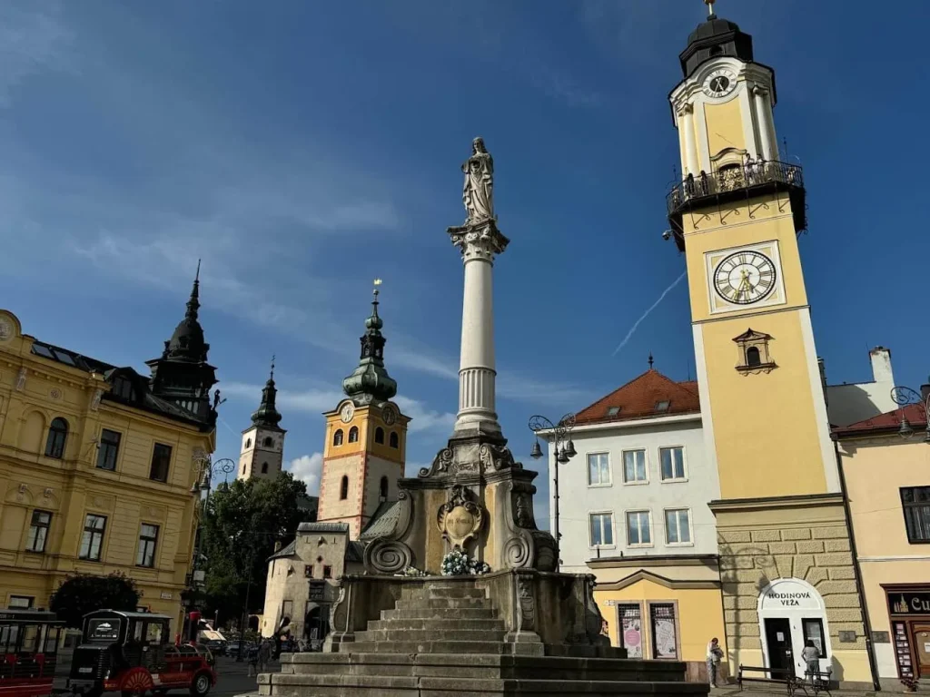 A Slovak town square with the leaning Clock Tower on the right, the Marian column at its centre and church towers behind.