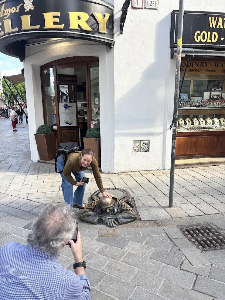 A visitor bending to touch Čumil, the bronze sewer-worker statue in Bratislava’s Old Town, while a companion photographs her.