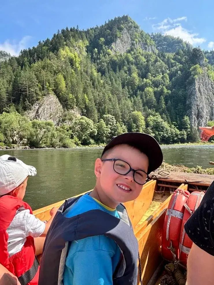 A boy in a life jacket on a wooden raft on the Dunajec, the forested limestone cliffs of the Pieniny behind him.