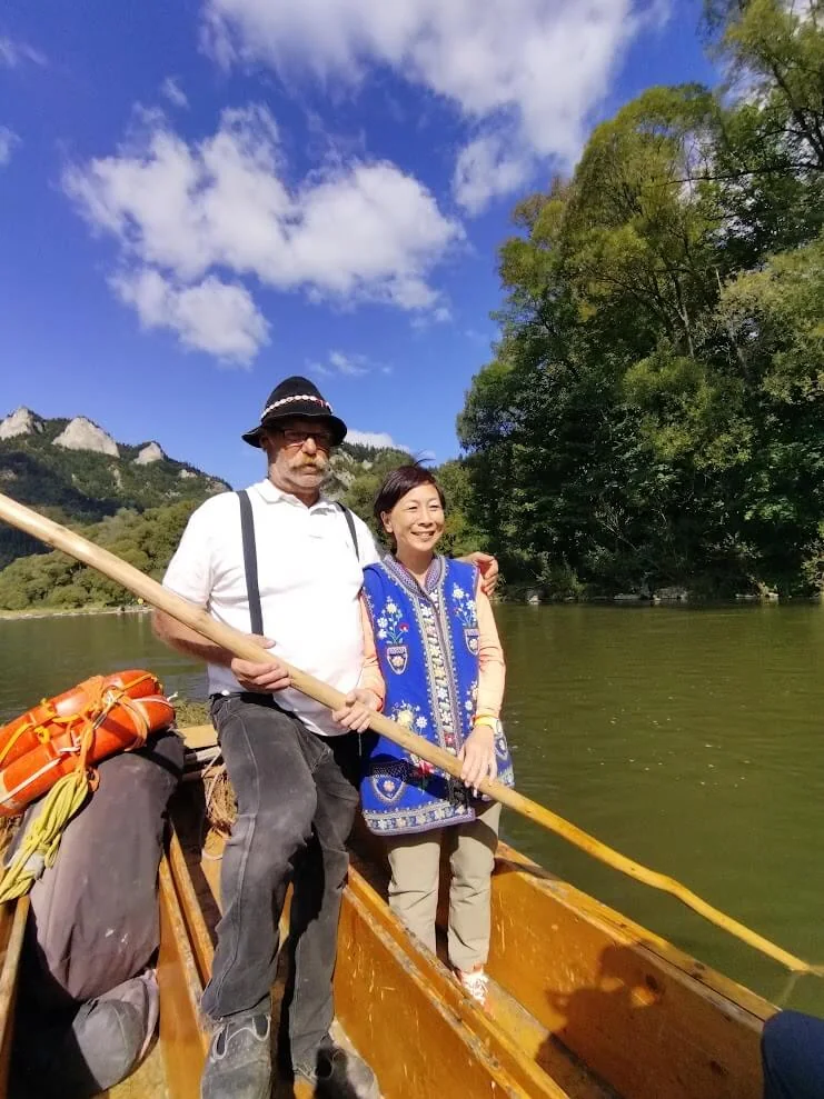 A raftsman in traditional dress poling a wooden raft on the Dunajec, a guest standing beside him and the limestone peaks of the Pieniny behind.