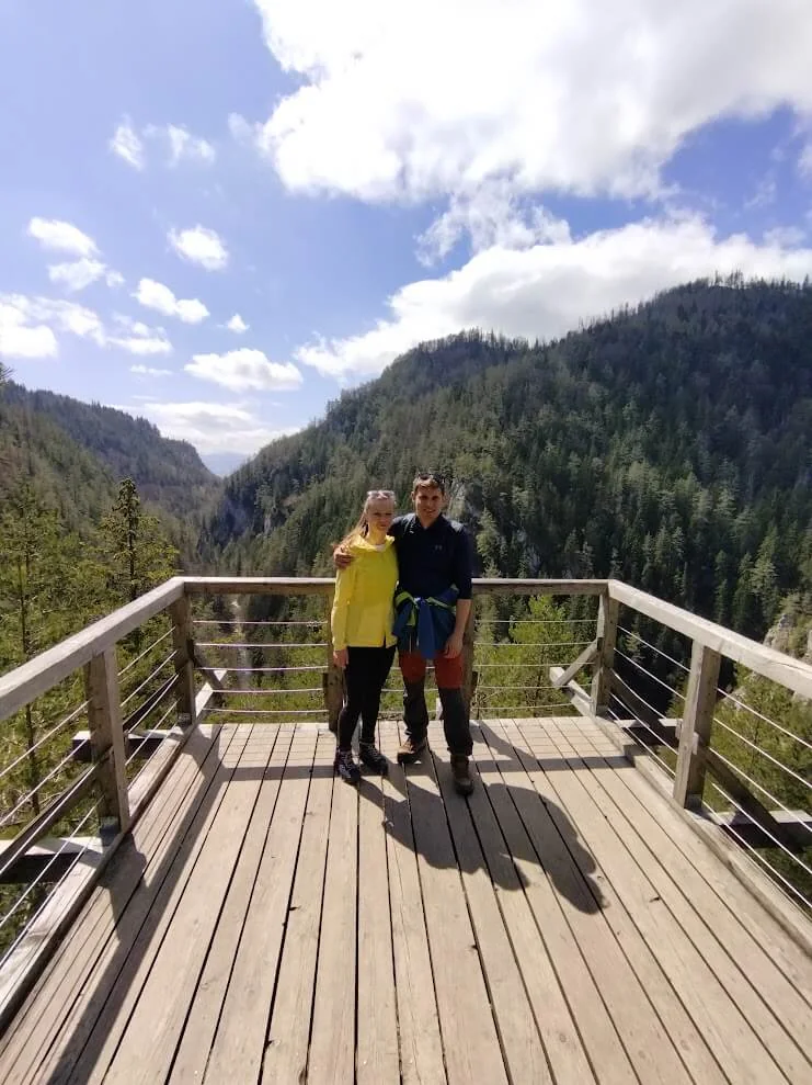Two visitors on a wooden viewing platform above a forested gorge in Slovakia, limestone cliffs rising on both sides.