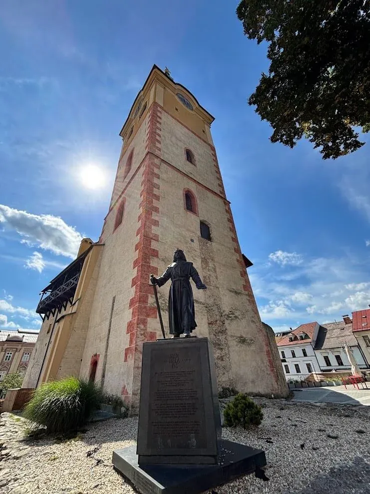 A tall stone bell tower in a Slovak mining town, with a bronze statue of King Béla IV on a plinth in front of it.