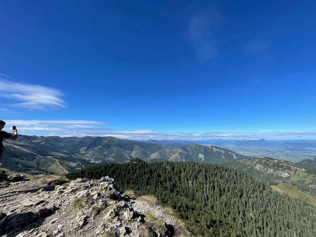 A walker photographing the view from a stony summit in Slovakia, forested ridges and a broad valley spread out below.