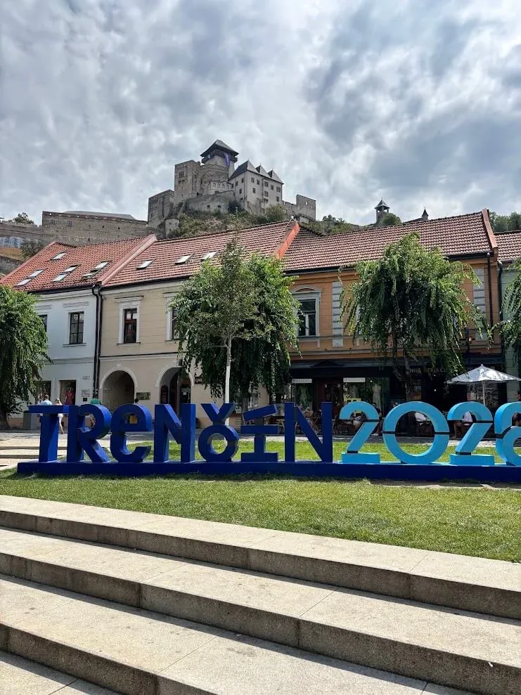 Trenčín’s main square with the TRENČÍN 2028 sign in blue letters and Trenčín Castle on the rock above the rooftops.