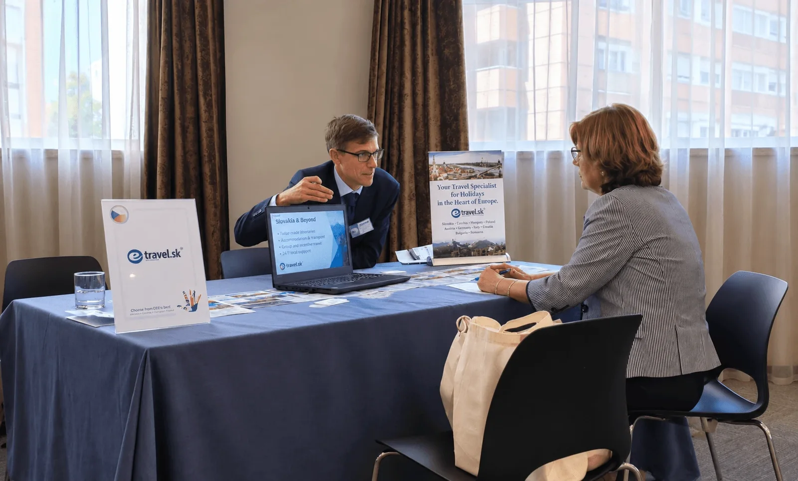 An e-travel.sk representative in conversation with a buyer across a table at a travel trade workshop, the company’s Slovakia display boards and a laptop between them.